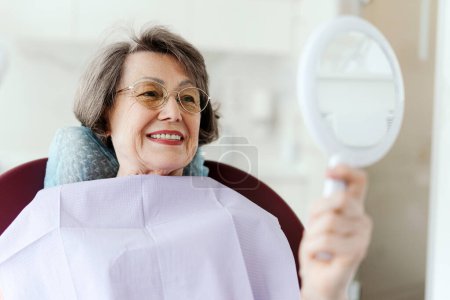 Smiling senior atractiva mujer paciente sosteniendo espejo mirando los dientes mientras está sentado en la silla dental en la odontología moderna. Concepto de cuidado dental, cuidado de la salud