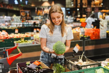 Smiling woman with blonde hair selects fresh savoy cabbage in a grocery store vegetable aisle, filling her cart with green, healthy options. Shopping concept