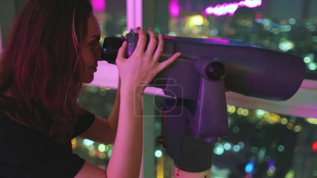 Foto de Mujer mirando en telescopio en la ciudad nocturna. El turista disfruta del mirador Bangkok Baiyoke Tower Sky. Centro de paisaje urbano colorido en el centro, vistas de luces nocturnas desde el último piso. Viajes, turismo - Imagen libre de derechos