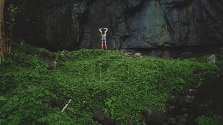 Woman enjoy jungle rock Bali waterfall landscape standing on green hill. Girl relax outdoor lifestyle travel on summer holiday vacation. Picturesque drone aerial flight. Dramatic dark toning back view