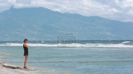 Photo for Lonley woman standing on ocean coast looking on waves crashing in windy weather. Young slim brunette relax on tropical island beach, dramatic moody scene. Travel, tourism, holiday. - Royalty Free Image