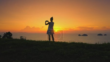 Silhouette femme contre le coucher de soleil vibrant, soleil couchant rayonnant. Champ herbeux, ciel orange, fille avec caméra capture nature beauté océan îles. Touriste féminine en vacances d'été. Lifestyle vacances
