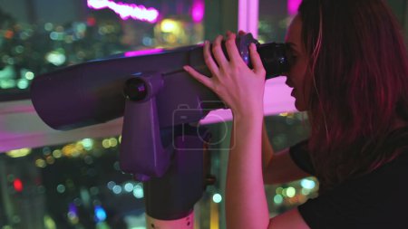 Téléchargez les photos : Femme heureuse regardant paysage urbain de nuit dans le télescope. Touriste bénéficie Bangkok Baiyoke Tower Sky belvédère. Centre-ville coloré au centre-ville, vue sur les veilleuses depuis le dernier étage. Voyages, tourisme, - en image libre de droit