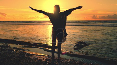 Woman raise hands up on sunset sandy sea beach, ocean waves in background. Female silhouette stand on ocean coast. Orange sky reflect in water. Relax, enjoy summer vacation. Back view
