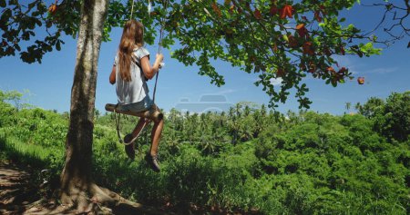 Una mujer está sentada en un columpio colgado de un árbol en Bali, específicamente en Ubuds Campuhan Ridge Walk. El columpio se balancea suavemente mientras disfruta del entorno sereno.