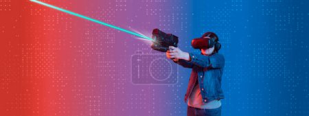 Téléchargez les photos : Young Hispanic woman wearing virtual reality goggles standing viewed from the front, holding plastic toy gun with both hands, shooting upwards, illuminated with red light against a virtual background - en image libre de droit