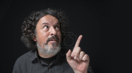 Téléchargez les photos : Portrait d'homme latin à la barbe blanche et aux cheveux bouclés noirs avec une expression drôle, pointant vers le haut avec sa main, portant une chemise noire sur fond noir - en image libre de droit