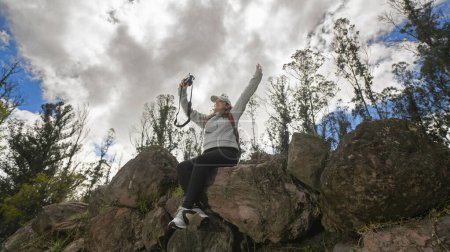 Téléchargez les photos : Belle jeune femme latino-américaine assise sur des pierres au sommet d'une montagne prenant un selfie avec sa petite caméra par une journée nuageuse - en image libre de droit