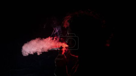 Téléchargez les photos : Portrait d'un homme adulte avec une cigarette à la main, soufflant de la fumée de sa bouche vue de profil en silhouette avec une lumière rouge illuminant son profil sur fond noir - en image libre de droit
