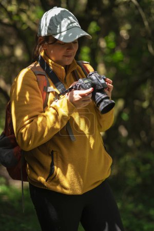 Téléchargez les photos : Belle femme latino-américaine marchant avec un sac à dos rouge et une casquette bleue revoyant les photos sur son appareil photo au milieu d'une forêt lors d'une journée ensoleillée - en image libre de droit