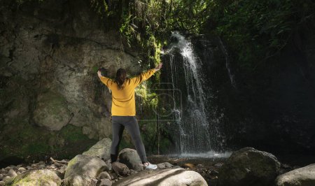 Téléchargez les photos : Belle femme latino-américaine vêtue d'une veste jaune et d'un pantalon noir debout à côté d'une cascade, les bras ouverts, au milieu d'une forêt par une journée ensoleillée - en image libre de droit