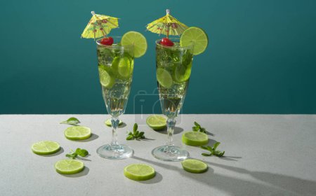 Téléchargez les photos : Deux cocktails en long verre remplis de tranches de glace et de citron garnis de petits parapluies jaunes, de cerises et de tranches de citron sur une table blanche sur fond turquoise - en image libre de droit