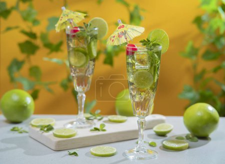 Téléchargez les photos : Deux cocktails en long verre remplis de tranches de glace et de citron garnis de petits parapluies jaunes, de cerises et de tranches de citron sur une planche de bois sur fond orange avec des feuilles vertes déconcentrées - en image libre de droit