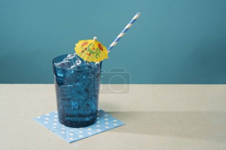 Téléchargez les photos : Cocktail en verre bleu rempli de glace avec petit parapluie jaune et paille sur serviette bleue sur une table blanche sur fond bleu - en image libre de droit