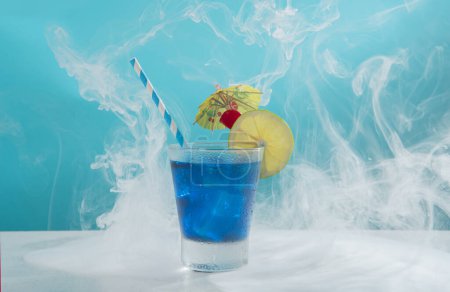 Téléchargez les photos : Cocktail sucré dans un verre froid rempli de glace garnie de tranches de citron, cerise, parapluie jaune et sorbet sur une table blanche sur fond bleu clair avec de la fumée - en image libre de droit