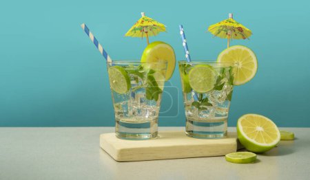 Téléchargez les photos : Deux cocktails en verre remplis de tranches de glace et de citron garnies de tranches de citron, de parapluies jaunes et de sorbets sur une planche de bois sur fond turquoise - en image libre de droit