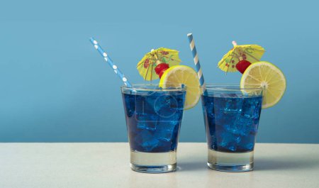 Téléchargez les photos : Deux cocktails bleus dans un verre rempli de glace garnie de tranches de citron, cerises, parapluies jaunes et sorbets sur une table blanche sur fond bleu - en image libre de droit