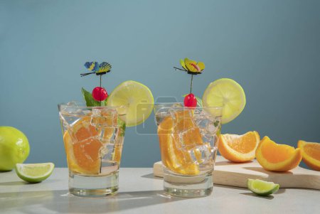 Téléchargez les photos : Deux cocktails en verre rempli de glace et de tranche d'orange garnie de tranche de citron, cerise et épinglette papillon sur une table blanche avec des morceaux d'orange et de citron sur fond bleu foncé - en image libre de droit