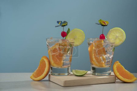 Téléchargez les photos : Deux cocktails en verre rempli de glace et de tranche d'orange garnie de tranche de citron, cerise et épinglette papillon sur une planche de bois avec des morceaux d'oranges et de citron sur la table sur fond bleu - en image libre de droit
