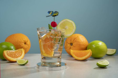 Téléchargez les photos : Cocktail en verre rempli de glace et de tranche d'orange garni de tranche de citron, cerise et épinglette papillon sur une table blanche avec des morceaux d'orange et de citron sur fond bleu foncé - en image libre de droit