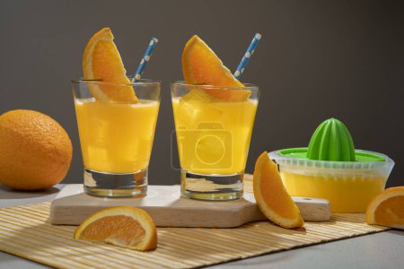 Téléchargez les photos : Deux cocktails jaunes en verre rempli de glace garnie d'une tranche d'orange et de sorbet sur une serviette en bambou avec des morceaux d'orange et un petit presse-jus sur fond gris foncé - en image libre de droit