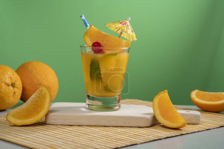 Téléchargez les photos : Cocktail jaune en verre rempli de feuilles de glace et de menthe garnies de tranches d'orange, cerise, sorbet et parapluie jaune sur une serviette en bambou avec des morceaux d'orange sur fond vert foncé - en image libre de droit