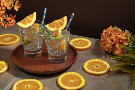 Téléchargez les photos : Deux cocktails dans un verre rempli de feuilles de glace et de menthe garnies d'une tranche d'orange et de sorbet sur une assiette en bois sur une table en céramique avec des tranches d'orange sur un fond rouge foncé - en image libre de droit