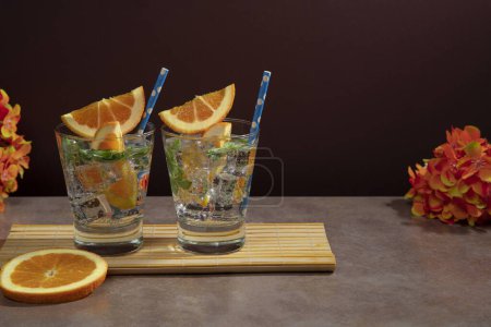 Téléchargez les photos : Deux cocktails en verre rempli de glace, feuilles de menthe garnies de tranches d'orange et sorbet sur une serviette en bambou sur une table en céramique avec une tranche d'orange et des fleurs rouges sur un fond rouge foncé - en image libre de droit