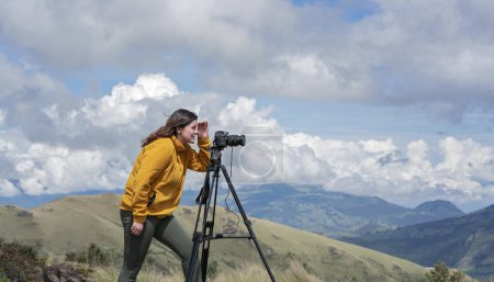 Téléchargez les photos : Belle jeune femme latino-américaine avec son appareil photo et son trépied prenant des photos de paysages au sommet d'une montagne lors d'une journée ensoleillée - en image libre de droit