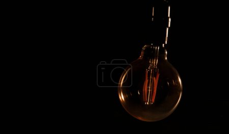 Téléchargez les photos : Ampoule en verre non éclairée sans électricité suspendue avec des reflets chauds sur fond noir - en image libre de droit