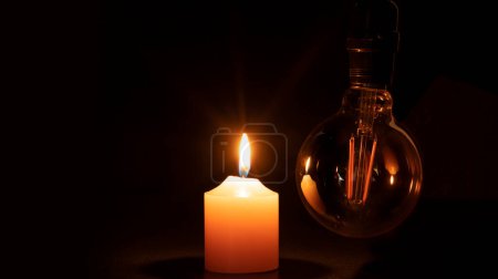 Téléchargez les photos : Concept de panne d'électricité, éteint l'ampoule sans électricité suspendue à côté d'une bougie allumée sur fond noir - en image libre de droit