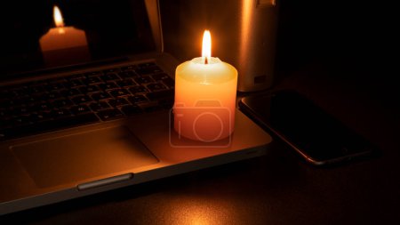 Téléchargez les photos : Blackout concept, Une bougie blanche allumée sur un ordinateur portable sur un fond noir - en image libre de droit