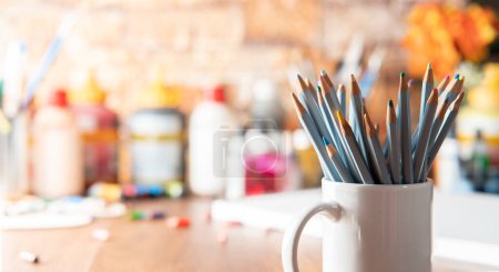 Téléchargez les photos : Tasse blanche avec des crayons de couleur au premier plan sur une table en bois avec des fournitures d'art en arrière-plan hors foyer - en image libre de droit