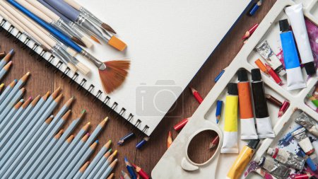 Téléchargez les photos : Vue du dessus de la table en bois avec crayons, tubes de peinture, fournitures d'art, variété de pinceaux sur papier blanc - en image libre de droit