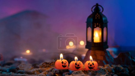 Téléchargez les photos : Fond d'Halloween, 3 bougies en forme de citrouille au premier plan, une vieille lampe sur un rocher contre un fond bleu flou avec des lumières jaunes et de la brume - en image libre de droit