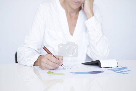Foto de Detalle de una mujer de mediana edad con una camisa blanca sentada en una mesa blanca, trabajando con documentos con gráficos y estadísticas financieras sosteniendo un bolígrafo en su mano derecha. - Imagen libre de derechos