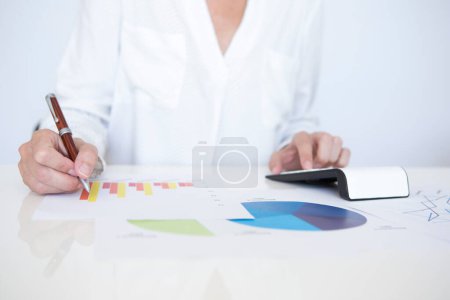 Foto de Detalle de una mujer de mediana edad con una camisa blanca sentada en una mesa blanca, trabajando con documentos con gráficos y estadísticas financieras sosteniendo un bolígrafo en su mano derecha. - Imagen libre de derechos