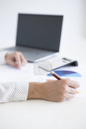 Foto de Manos de mujer de mediana edad en camisa blanca trabajando en estadísticas financieras con su computadora portátil y calculadora - Imagen libre de derechos