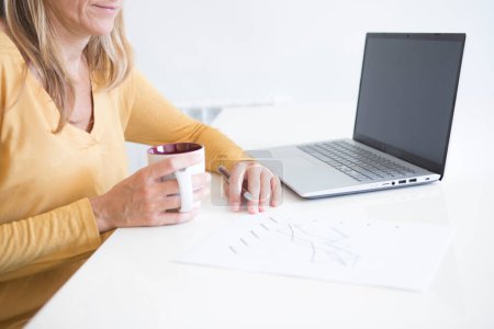 Foto de Manos de una mujer con camisa amarilla mostaza bebiendo una taza de té mientras trabaja con su móvil y su computadora en finanzas - Imagen libre de derechos