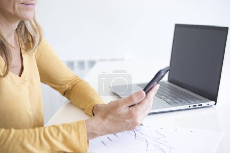 Foto de Manos de una mujer con camisa amarilla mostaza sosteniendo su teléfono móvil mientras analiza estadísticas financieras y gráficos - Imagen libre de derechos