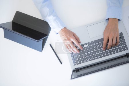 Foto de Manos de mujer de mediana edad en camisa de vestir azul trabajando con computadora portátil y tableta en escritorio blanco - Imagen libre de derechos