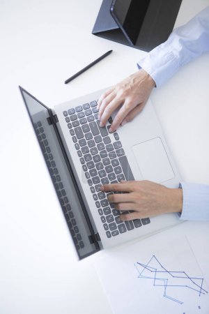 Foto de Vista aérea de las manos de la mujer de mediana edad en camisa de vestir azul trabajando con el ordenador portátil en el escritorio blanco - Imagen libre de derechos