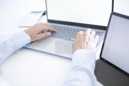 Foto de Manos de mujer de mediana edad en camisa de vestir azul trabajando con computadora portátil y tableta en escritorio blanco - Imagen libre de derechos
