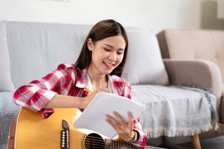 Junge Frau komponiert Song und schreibt Text auf Notizbuch, während sie die Gitarre auf die Beine stellt und zu Hause im Wohnzimmer auf dem Boden sitzt.