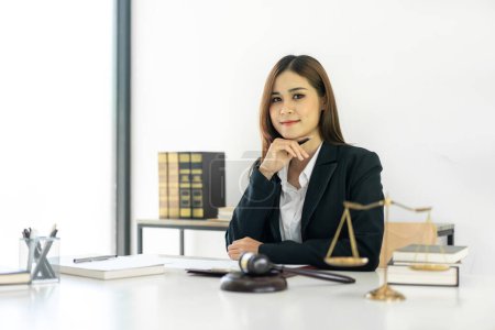 Photo for Business lawyer woman reading law book with contract document while touching chin on hand to looking at camera and working on the table with brass scales and justice hammer in law firm. - Royalty Free Image