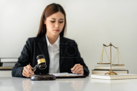 Photo for Business lawyer woman holding justice hammer and reading business contract to writing information on document while working on the table with brass scales in law firm office. - Royalty Free Image
