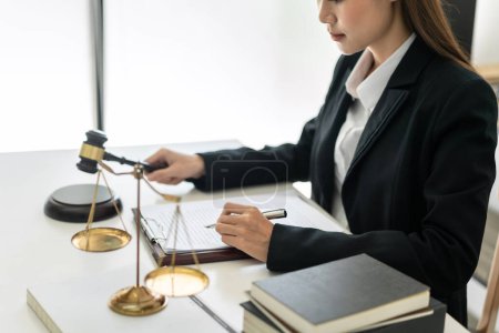 Photo for Business lawyer woman holding justice hammer and reading business contract to writing information on document while working on the table with brass scales in law firm office. - Royalty Free Image
