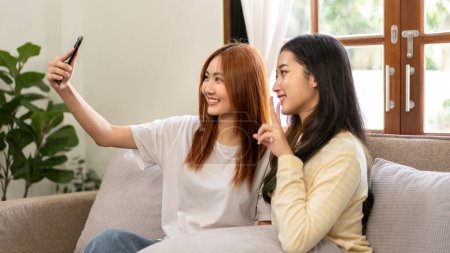 Photo for Young woman lesbian couple making v sign and smiling to taking photo selfie on smartphone while spending time to relaxation together on big comfortable sofa at home. - Royalty Free Image
