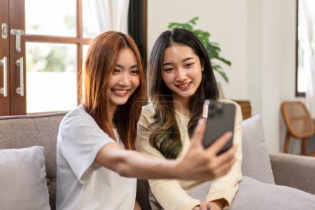 Téléchargez les photos : Jeune femme lesbienne couple souriant et prenant photo selfie sur smartphone tout en passant du temps à se détendre ensemble sur un grand canapé confortable à la maison. - en image libre de droit