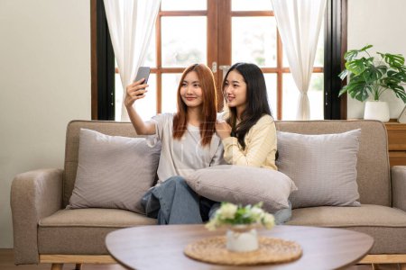 Photo for Young woman lesbian couple smiling and taking photo selfie on smartphone while spending time to relaxation together on big comfortable sofa at home. - Royalty Free Image